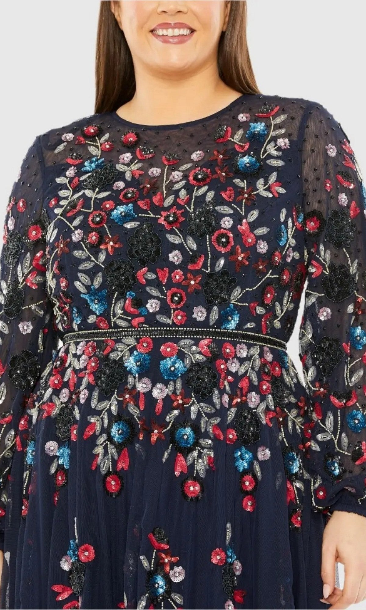A person wearing the Mac Duggal 94050 Plus-Size Cocktail Dress with Sleeves in navy blue, featuring colorful floral beadwork and sheer sleeves, smiles while facing forward.